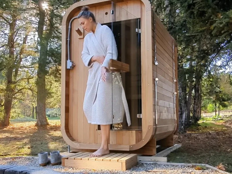Modern cedar sauna cabin in a forest setting with a guest on the entry deck