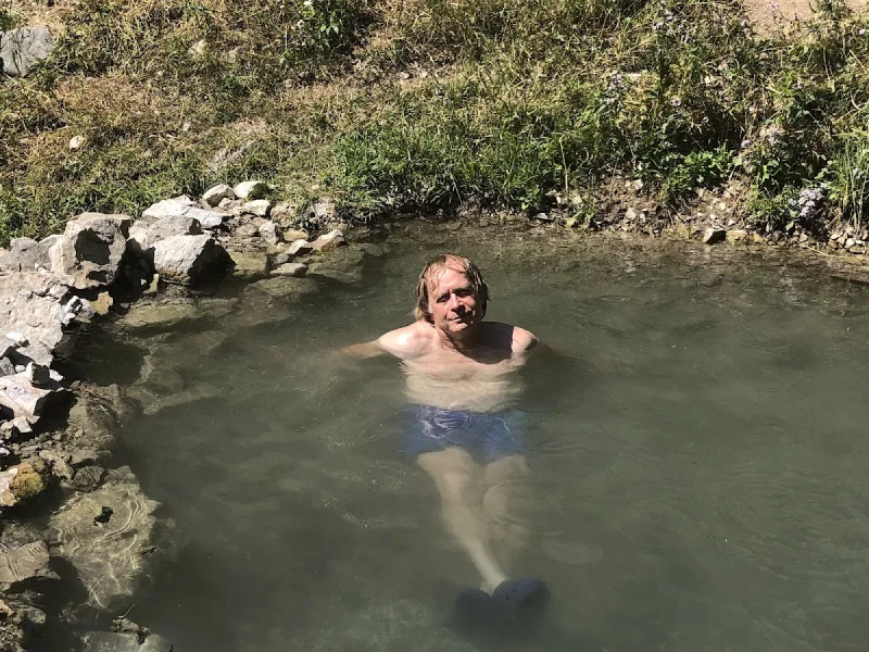 Man soaking in a rustic outdoor hot spring pool beside grassy banks