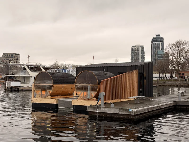 Floating barrel saunas beside a dock with city skyline and calm water