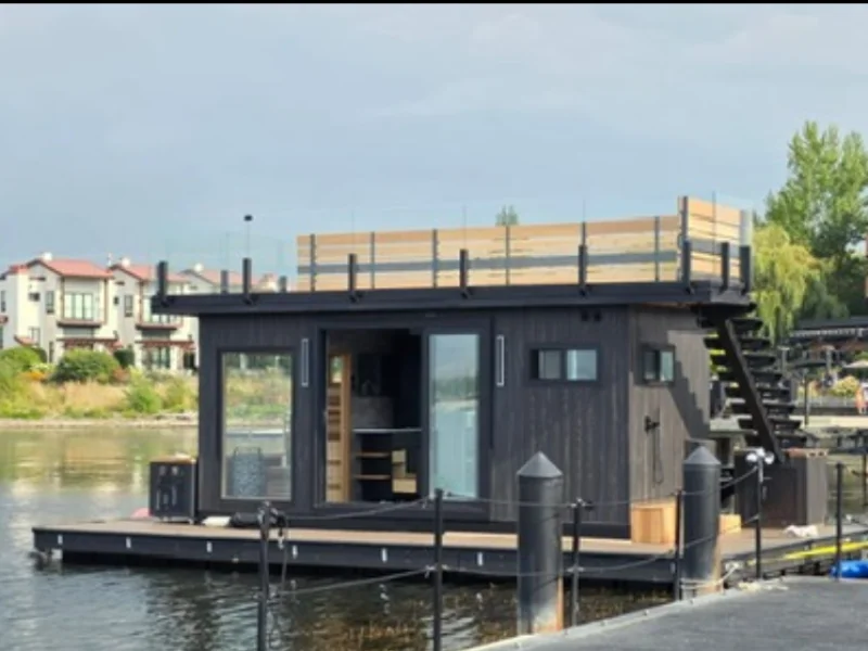 Floating sauna docked near waterfront homes with rooftop deck and entry stairs