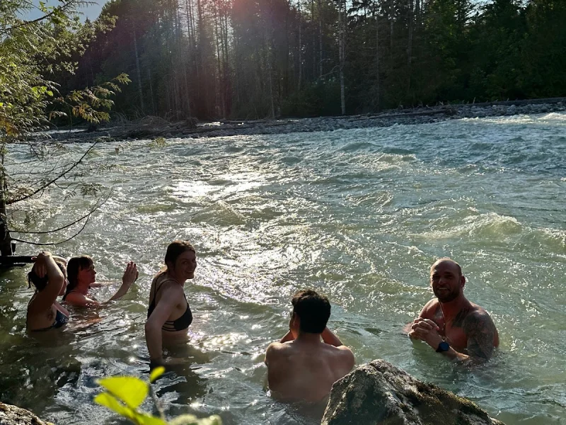 Guests relaxing in a cold river beside forested mountains at sunset